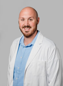 Professional headshot of a male Optima Medical provider wearing a white lab coat and light blue collared shirt, smiling against a neutral gray background.