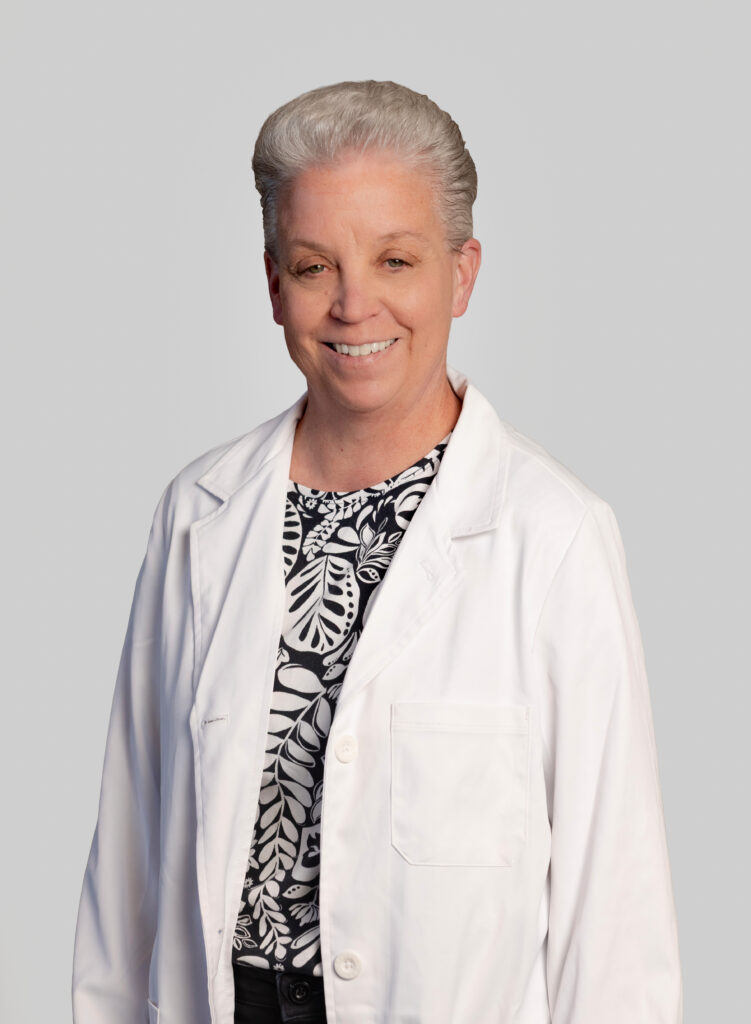 Female medical provider wearing a white lab coat, smiling, posed against a light gray background.